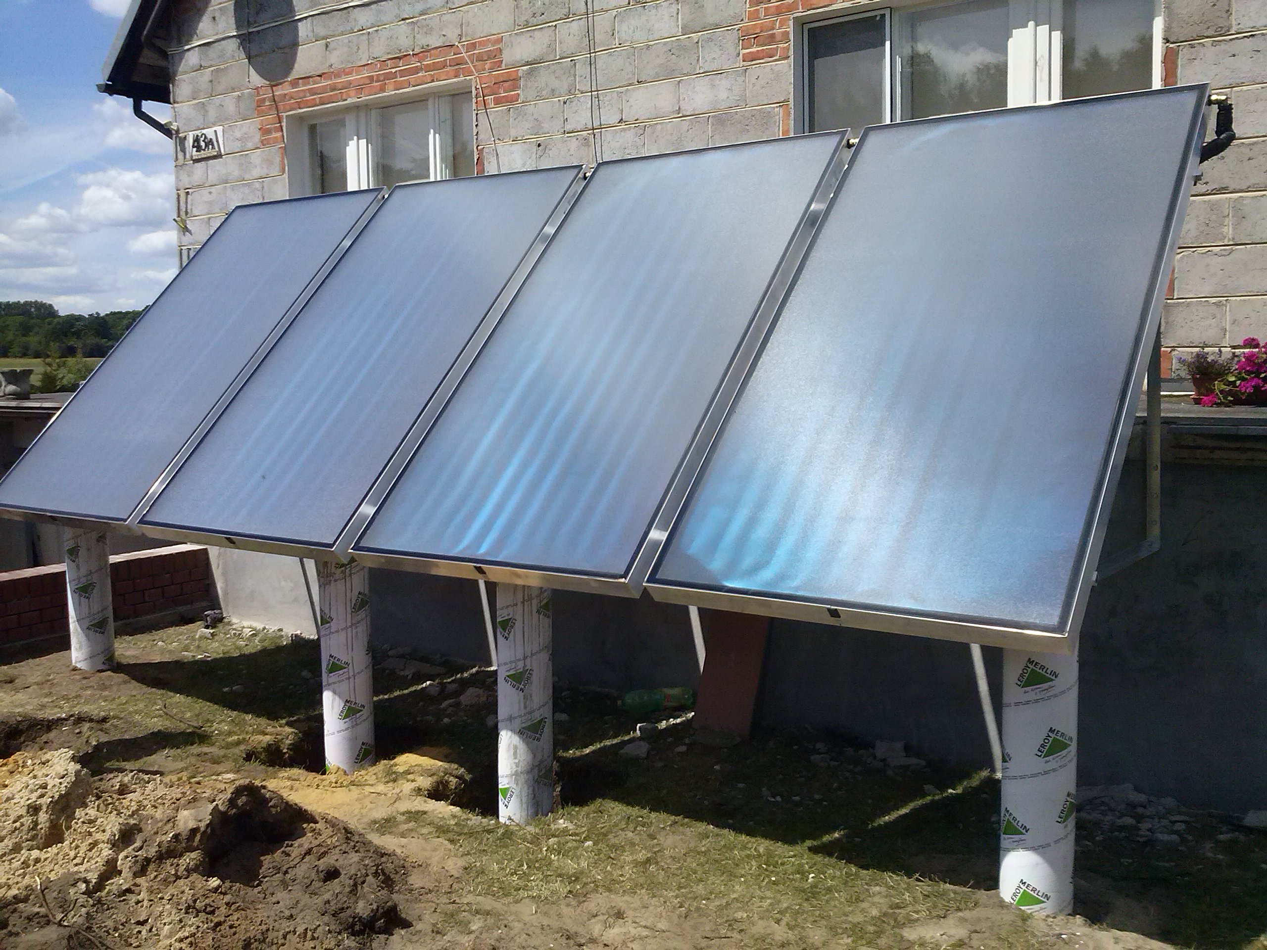 Cztery panele kolektorów słonecznych z metalową ramą, zamontowane na rurach osłonowych z nadrukiem marki budowlanej, ustawione przed budynkiem z cegły i pustaków w słoneczny dzień.