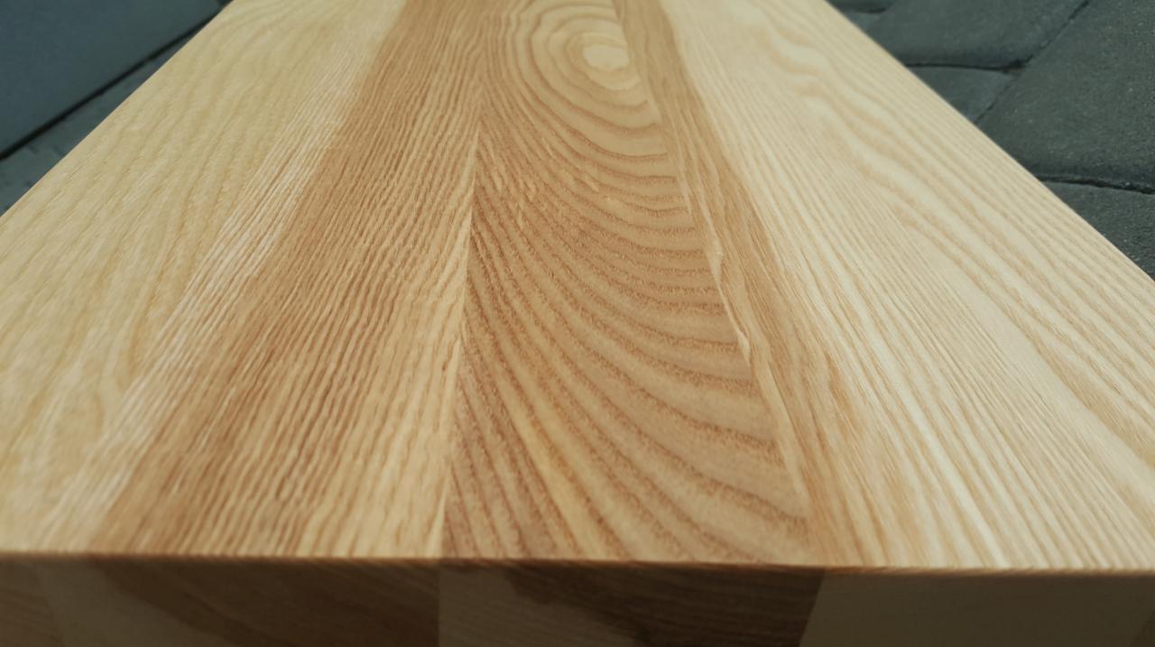 Jasna, drewniana deska o wyraźnym usłojeniu, sfotografowana pod kątem, ukazująca strukturę drewna i krawędź blatu.