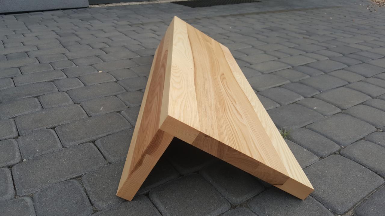 Kątowy element z drewna jesionowego, prawdopodobnie fragment schodów, leżący na szarej kostce brukowej.