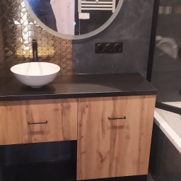 Teta Piotr Jóźwiak - Łazienkowa szafka pod umywalkę z drewnopodobnymi frontami i czarnym blatem, okrągłe lustro z podświetleniem LED i odbiciem osoby robiącej zdjęcie, złota mozaika na ścianie.