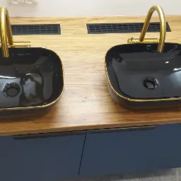 Dwie czarne, kwadratowe umywalki z złotymi krawędziami i złotymi bateriami na drewnianym blacie z niebieską szafką pod spodem.