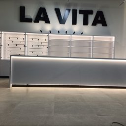 Teta Piotr Jóźwiak - Białe szafy na wymiar z podświetleniem LED i napisem 'LA VITA' na ścianie, widok z niskiego kąta.