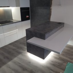 Teta Piotr Jóźwiak - Nowoczesna kuchnia z białymi szafkami i szarym blatem, wyspa kuchenna z podświetleniem LED u dołu, widoczne niedokończone wykończenie ściany.