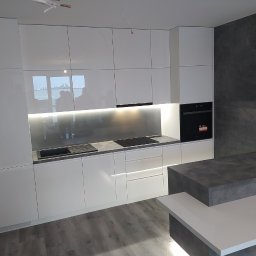 Teta Piotr Jóźwiak - Nowoczesna, biała kuchnia z połyskiem, podświetlane szafki wiszące, szary blat i wyspa kuchenna z elementami w kolorze betonu, widoczne niedokończone wykończenie ściany.