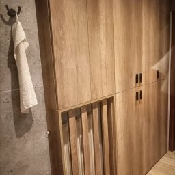 Teta Piotr Jóźwiak - Drewniana szafa z lamelami w łazience, obok wieszak z ręcznikiem na szarej ścianie.