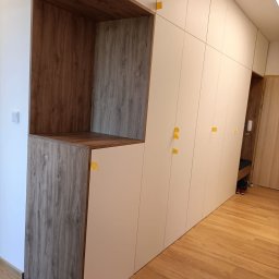 Teta Piotr Jóźwiak - Biała szafa wnękowa z drewnianymi akcentami i otwartą półką, zabezpieczona żółtymi taśmami montażowymi, na drewnianej podłodze.