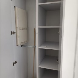 Teta Piotr Jóźwiak - Wnętrze białej, częściowo otwartej szafy z półkami i drążkiem na ubrania, widoczny również wystający z boku grzejnik i oparta o ścianę zmiotka.