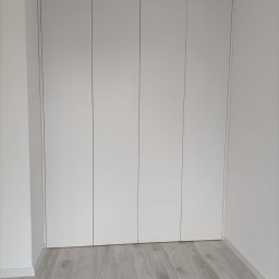 Teta Piotr Jóźwiak - Biała szafa wnękowa z czterema drzwiami bez klamek, na podłodze panele w odcieniach szarości, minimalistyczny design.