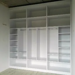 Biała, otwarta szafa wnękowa z półkami i drążkami na ubrania, w surowym wnętrzu z betonowym sufitem i jasną podłogą z płytek imitujących drewno.