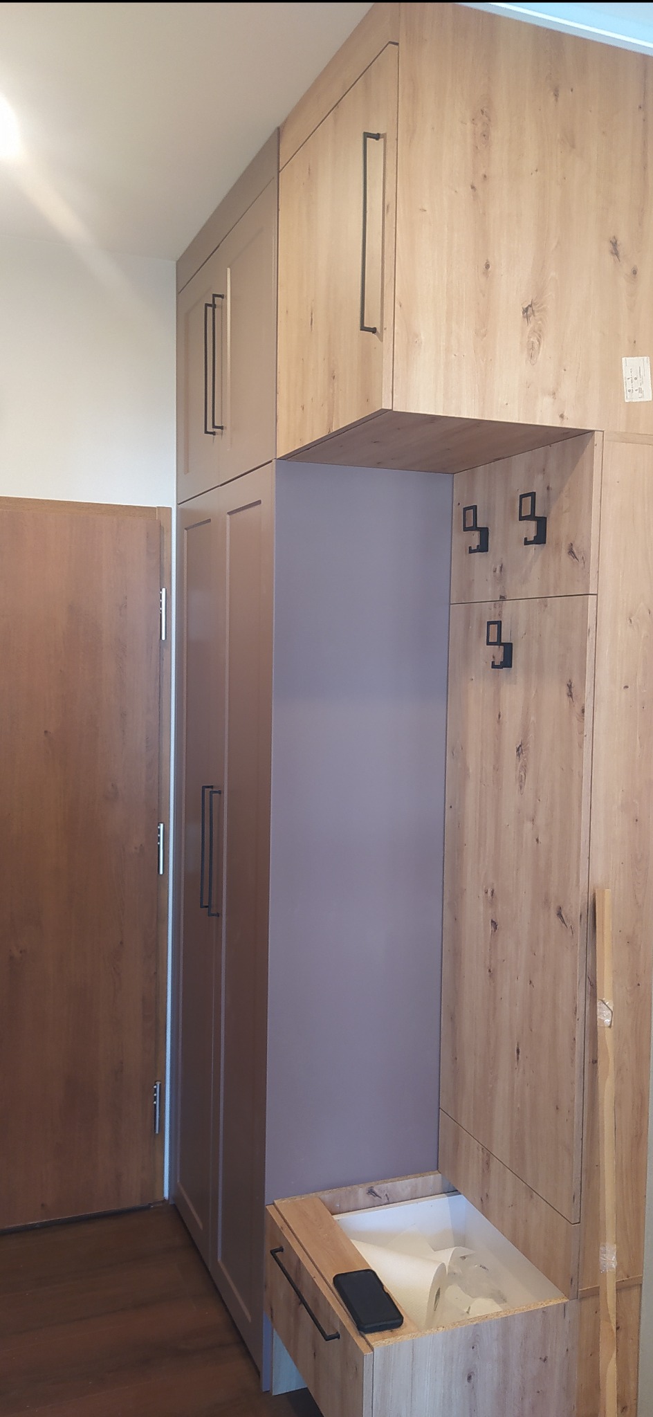 Szafa wnękowa z drewnianymi frontami, czarnymi uchwytami i wieszakami, z otwartą wnęką i szufladą, widoczny telefon na blacie szuflady.