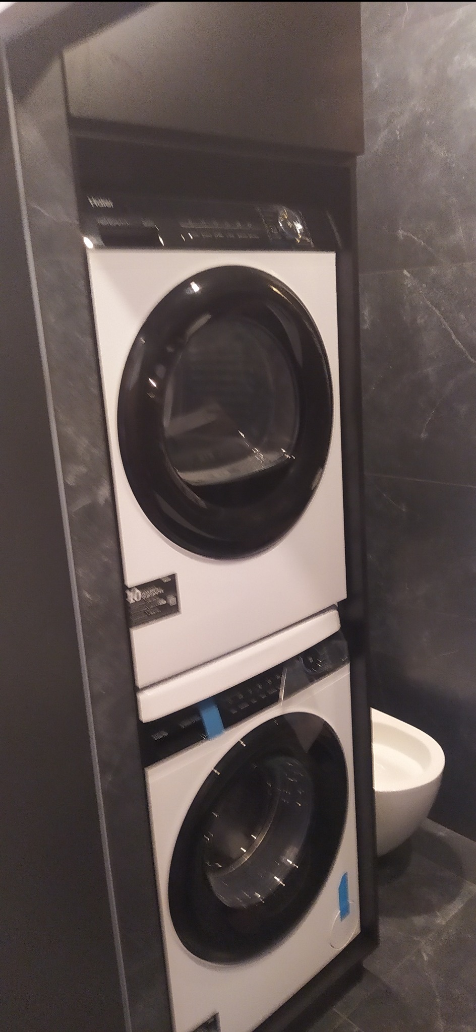 Zestaw pralko-suszarki Haier w zabudowie meblowej w łazience z szarą marmurową ścianą i białą umywalką.