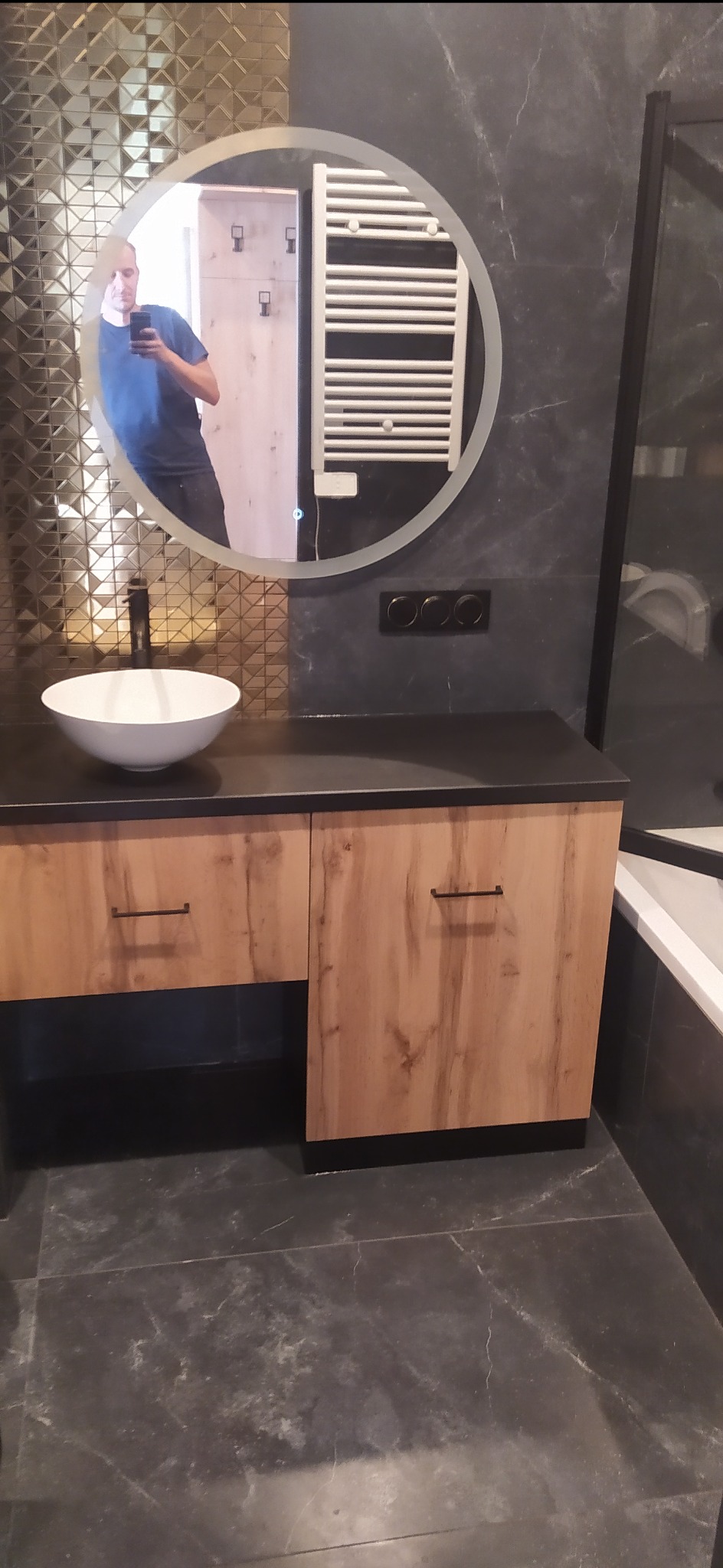 Łazienkowa szafka pod umywalkę z drewnopodobnymi frontami i czarnym blatem, okrągłe lustro z podświetleniem LED i odbiciem osoby robiącej zdjęcie, złota mozaika na ścianie.