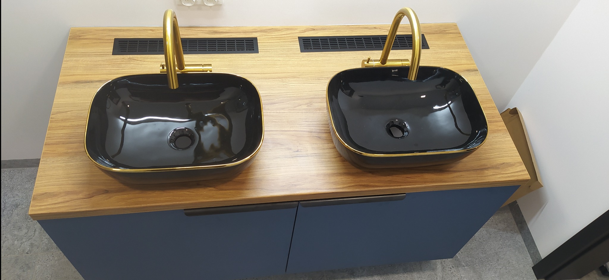 Dwie czarne, kwadratowe umywalki z złotymi krawędziami i złotymi bateriami na drewnianym blacie z niebieską szafką pod spodem.
