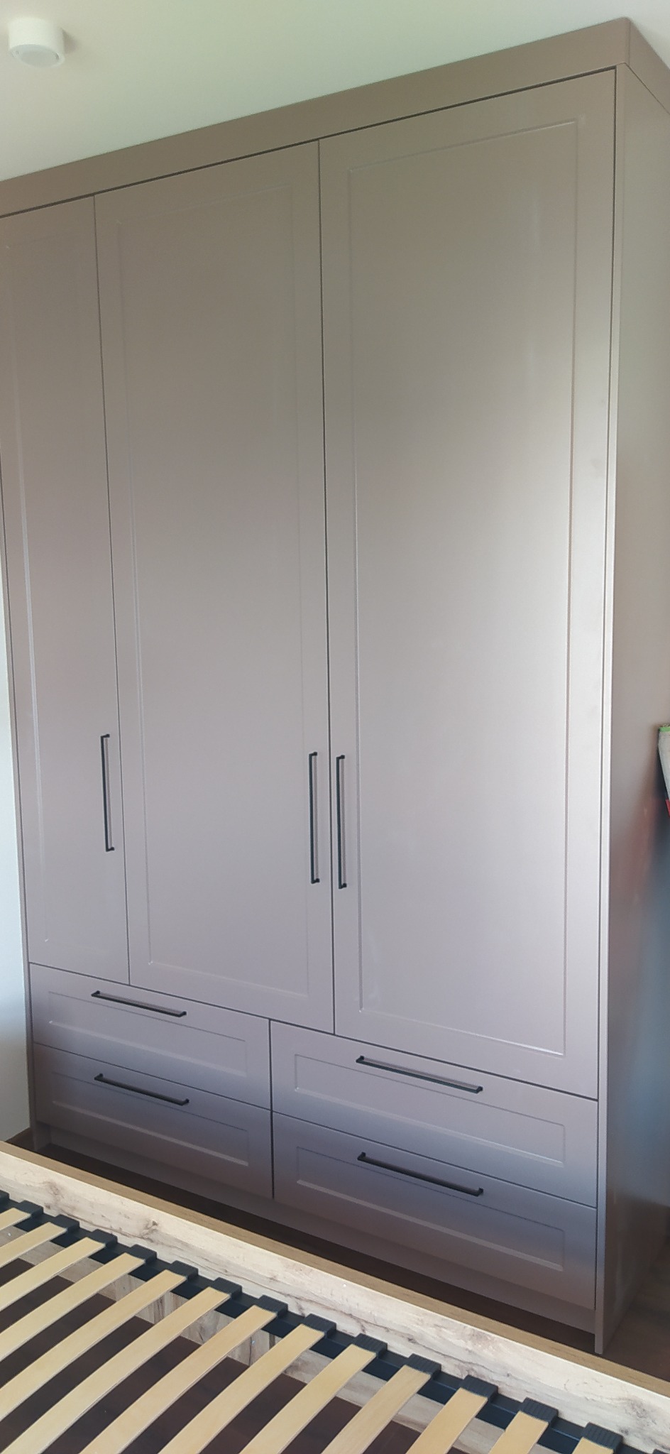 Szara szafa wnękowa z czterema drzwiami i czterema szufladami z czarnymi uchwytami, widoczny fragment ramy łóżka.