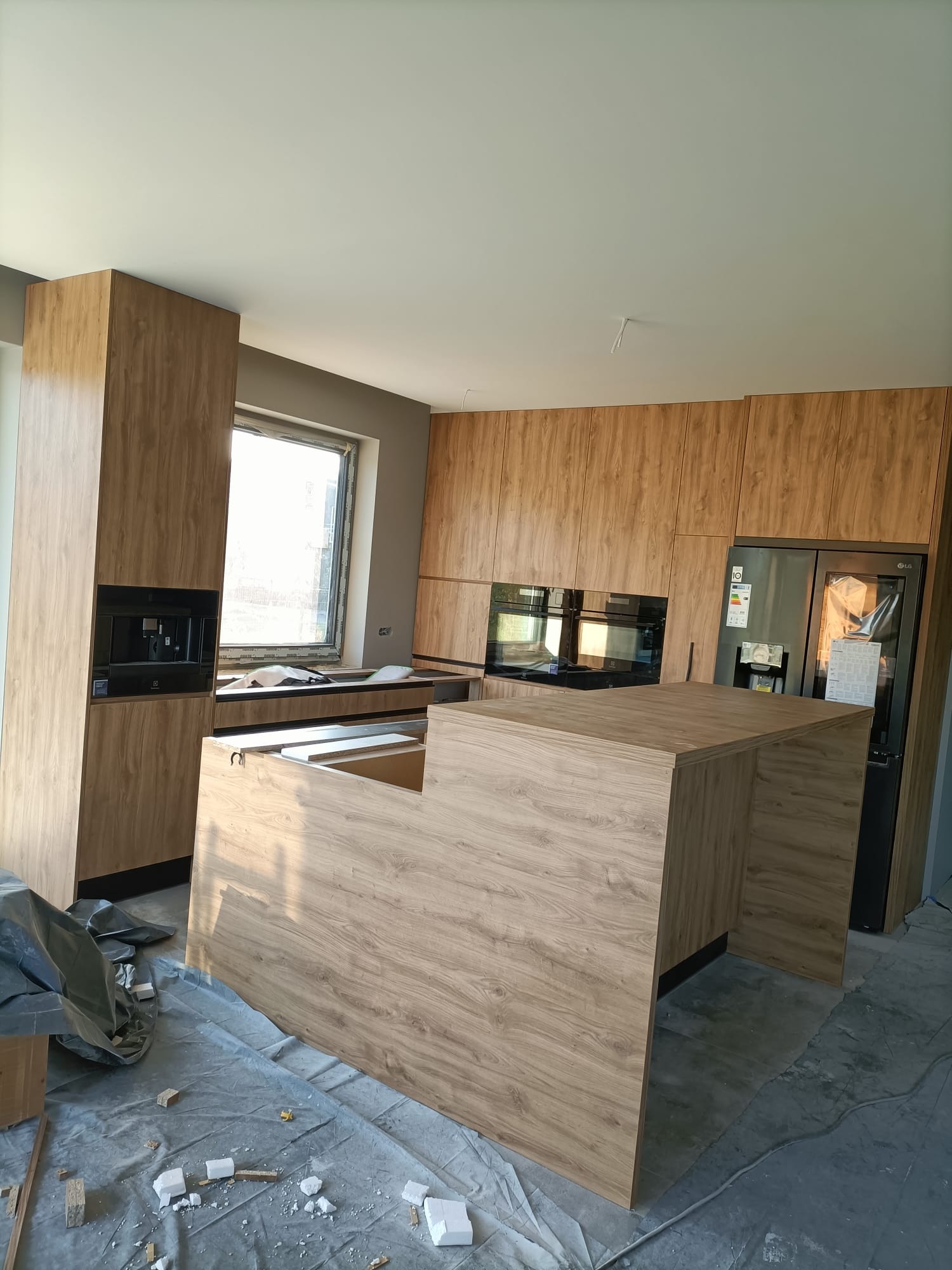 Montaż kuchni: wysoka zabudowa z wbudowanym ekspresem do kawy, szafki wiszące, wyspa kuchenna z blatem o wzorze drewna, lodówka side-by-side, widoczne zabezpieczenia podłogi.