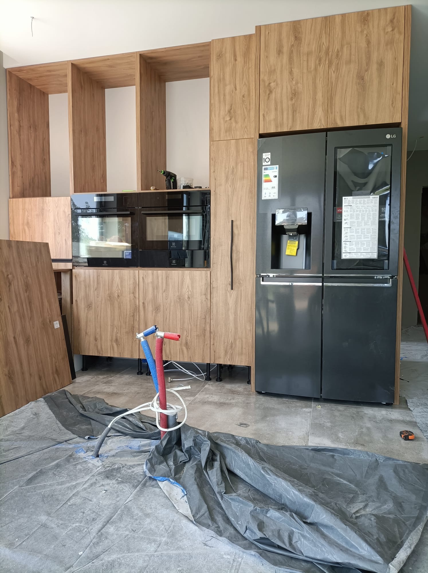 Częściowo zmontowana kuchnia z drewnianymi frontami i czarnym sprzętem AGD, w tym lodówka LG, widoczne rury instalacyjne i folia ochronna na podłodze.