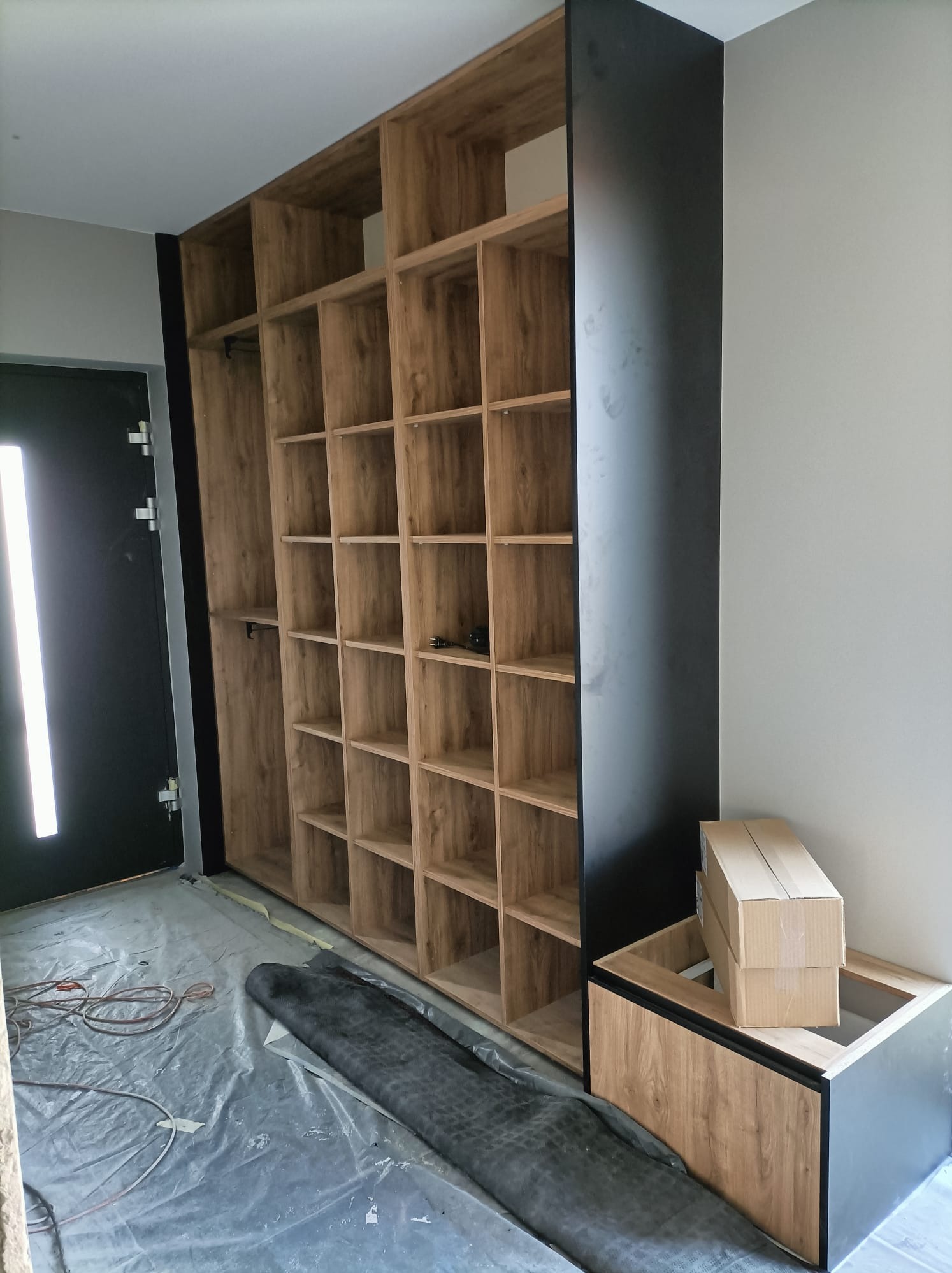 Montaż szafy wnękowej z drewnianymi półkami i czarnymi bokami; widoczne kartony i folia ochronna na podłodze.