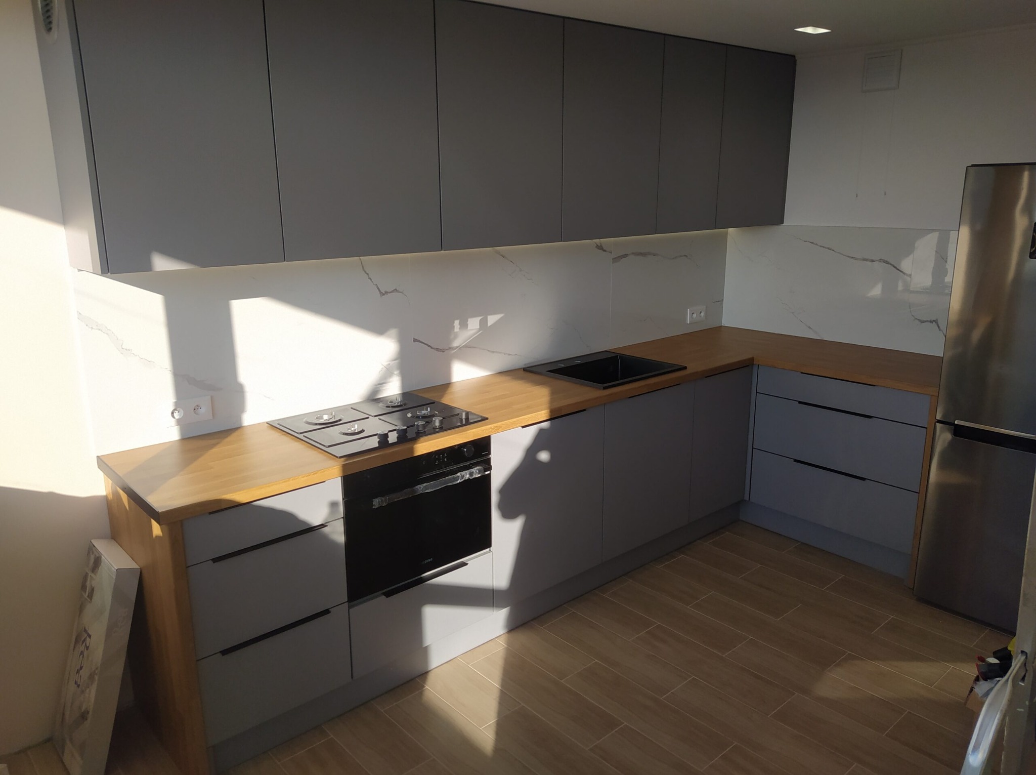 Szara zabudowa kuchenna z drewnianym blatem i marmurowym panelem ściennym, oświetlenie LED pod szafkami, płyta grzewcza i piekarnik w zabudowie, widok na podłogę z płytek imitujących drewno.