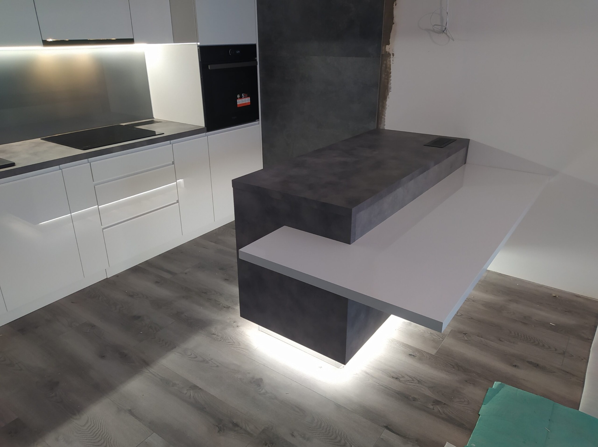 Nowoczesna kuchnia z białymi szafkami i szarym blatem, wyspa kuchenna z podświetleniem LED u dołu, widoczne niedokończone wykończenie ściany.