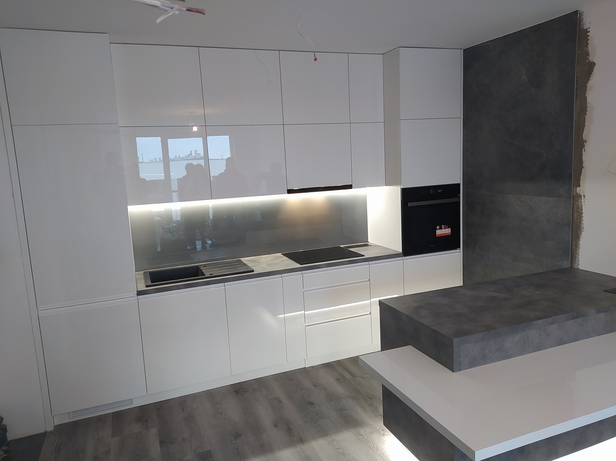 Nowoczesna, biała kuchnia z połyskiem, podświetlane szafki wiszące, szary blat i wyspa kuchenna z elementami w kolorze betonu, widoczne niedokończone wykończenie ściany.