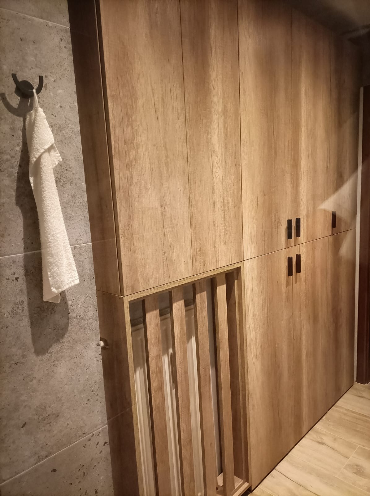 Drewniana szafa z lamelami w łazience, obok wieszak z ręcznikiem na szarej ścianie.