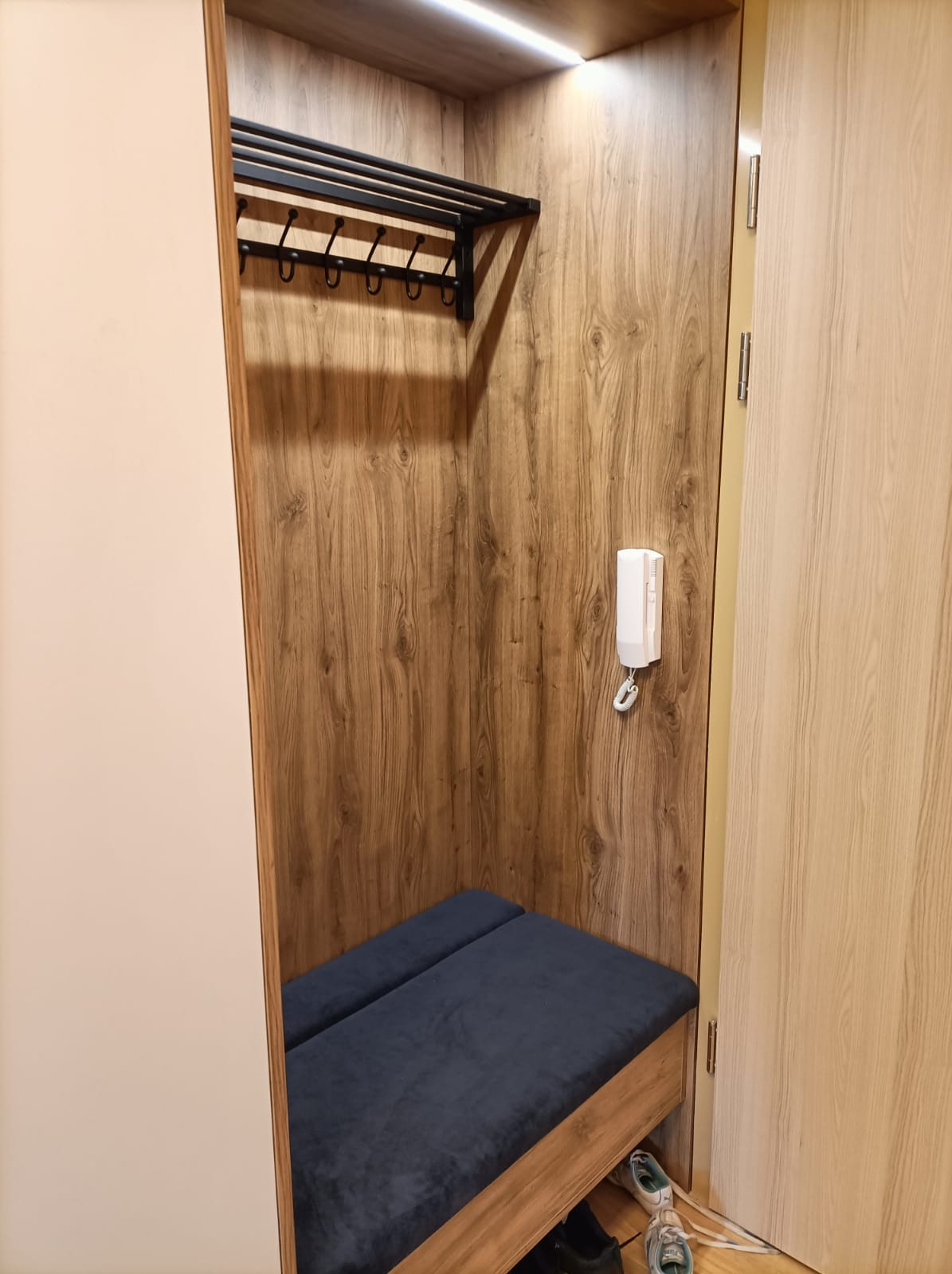 Wnęka garderoby z drewnianym wykończeniem, wyposażona w wieszak na ubrania, tapicerowane siedzisko i domofon na ścianie.
