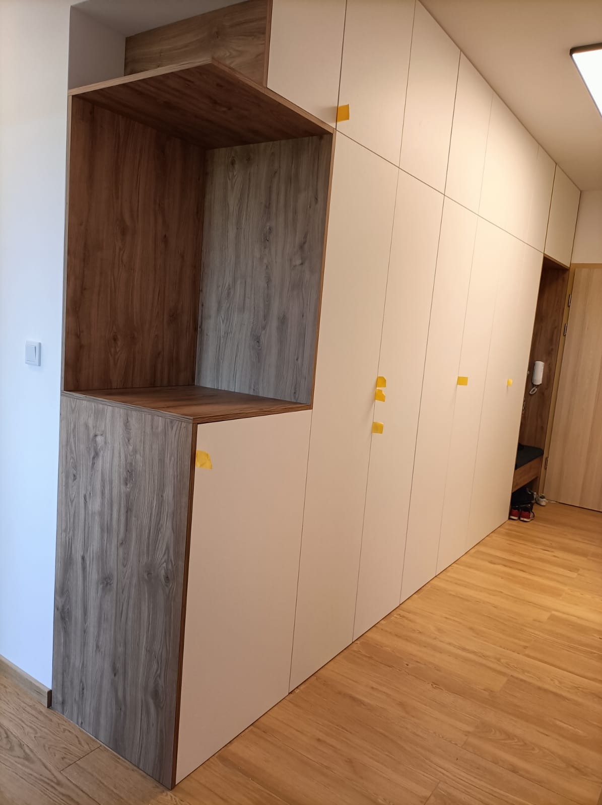 Biała szafa wnękowa z drewnianymi akcentami i otwartą półką, zabezpieczona żółtymi taśmami montażowymi, na drewnianej podłodze.