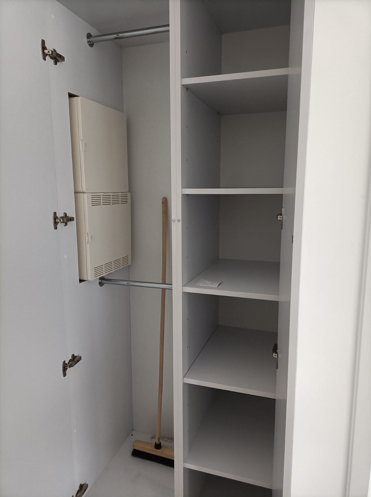Wnętrze białej, częściowo otwartej szafy z półkami i drążkiem na ubrania, widoczny również wystający z boku grzejnik i oparta o ścianę zmiotka.