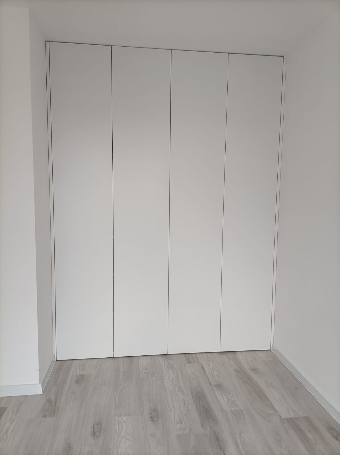 Biała szafa wnękowa z czterema drzwiami bez klamek, na podłodze panele w odcieniach szarości, minimalistyczny design.