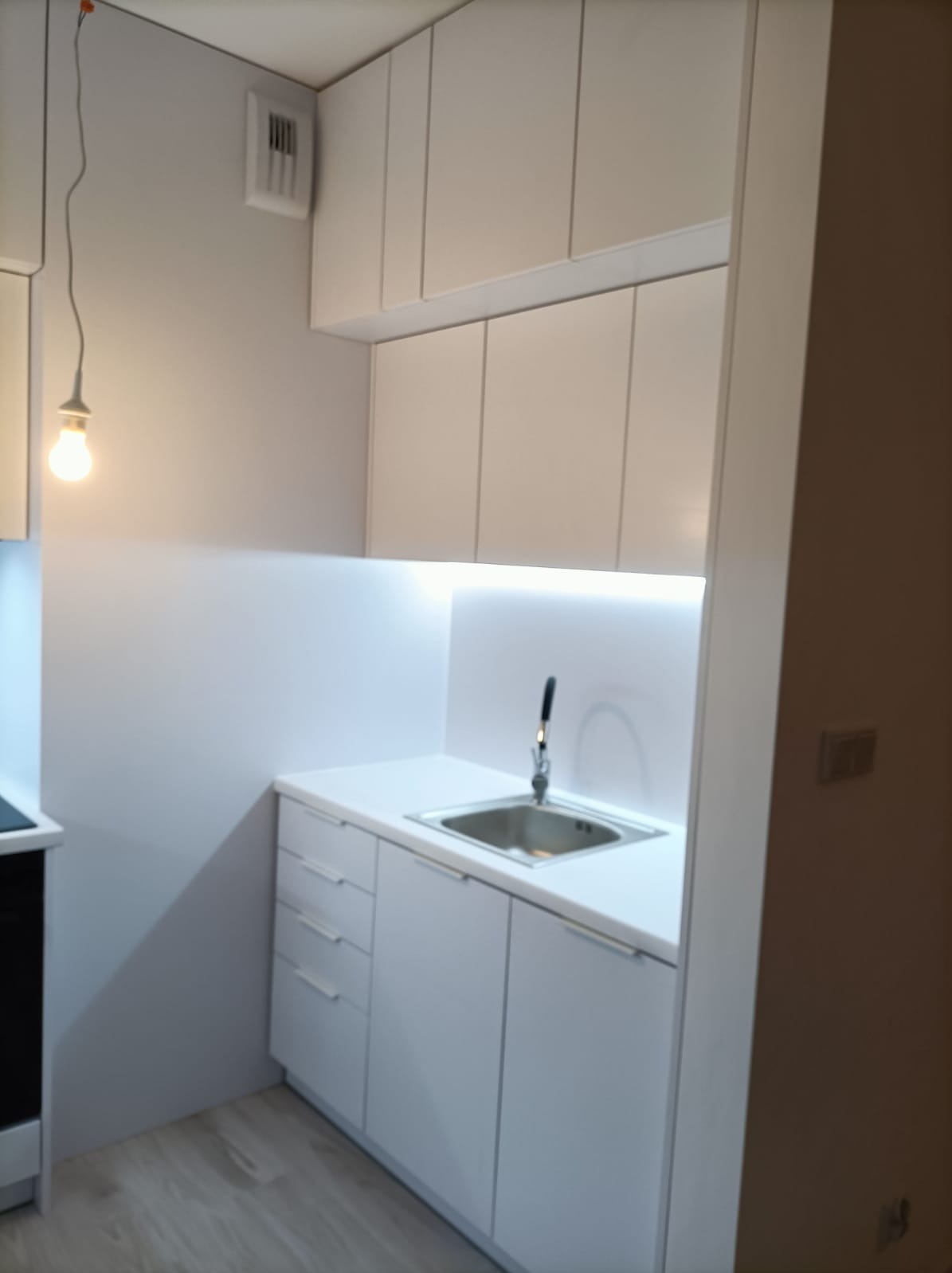 Nowoczesna, minimalistyczna kuchnia z białymi frontami szafek, oświetleniem LED podszafkowym i wiszącą żarówką na kablu.