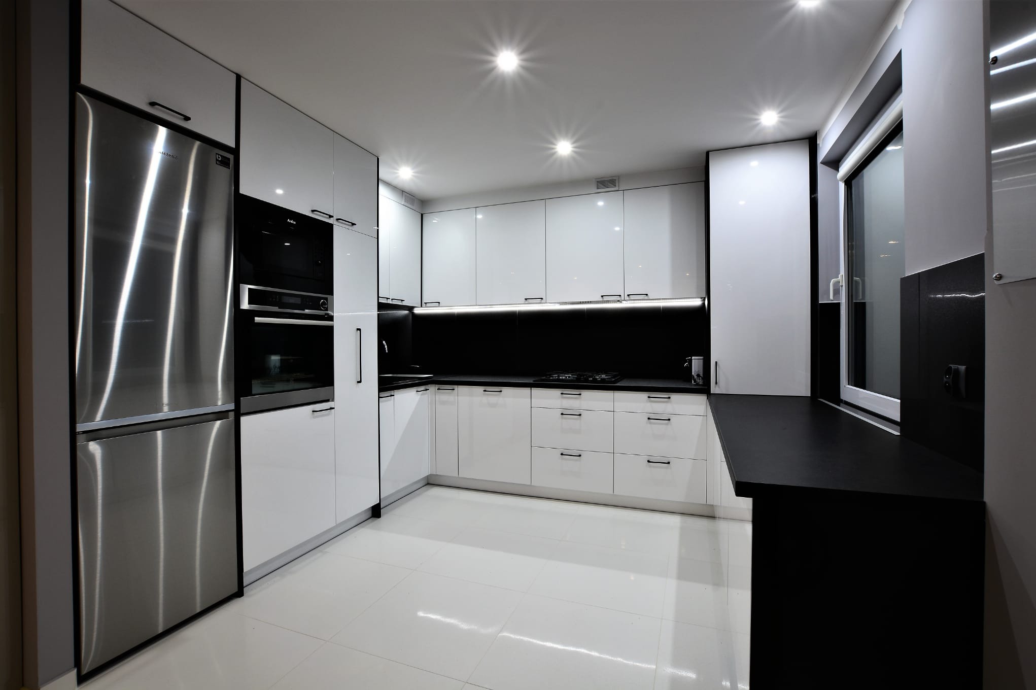 Nowoczesna kuchnia na wymiar z białymi, lakierowanymi frontami, czarnym blatem i zabudową AGD ze stali nierdzewnej.