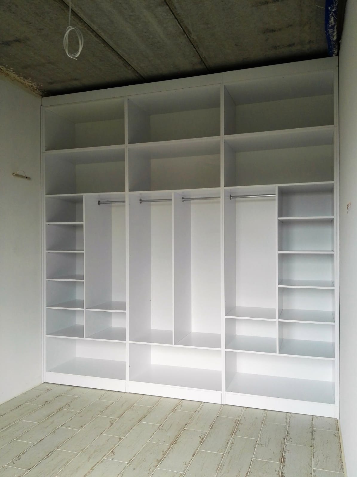 Biała, otwarta szafa wnękowa z półkami i drążkami na ubrania, w surowym wnętrzu z betonowym sufitem i jasną podłogą z płytek imitujących drewno.