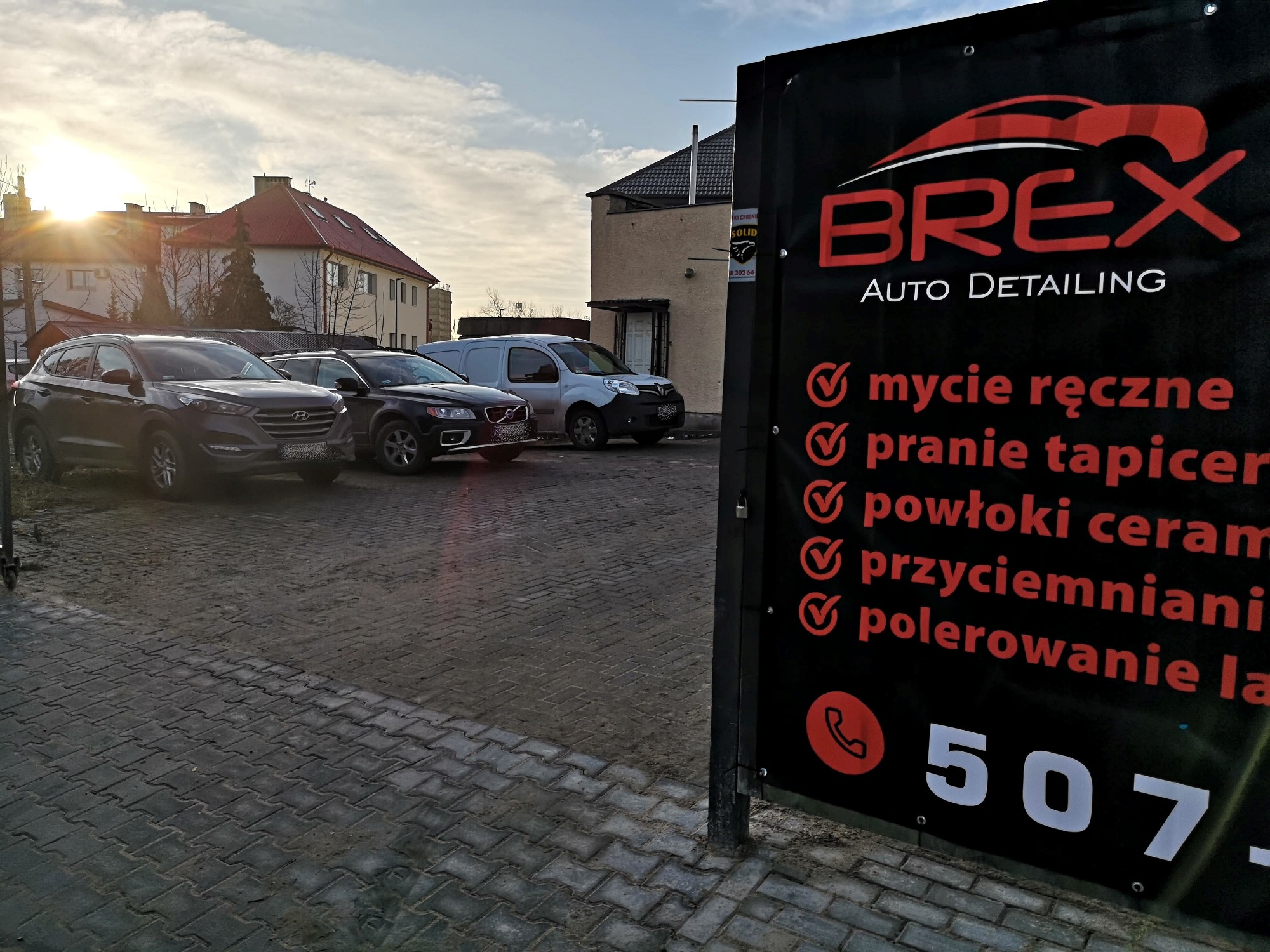 Czarny baner reklamowy firmy BREX Auto Detailing z listą usług (mycie ręczne, pranie tapicerki, powłoki ceramiczne, przyciemnianie, polerowanie lakieru) na tle zaparkowanych samochodów i budynku...