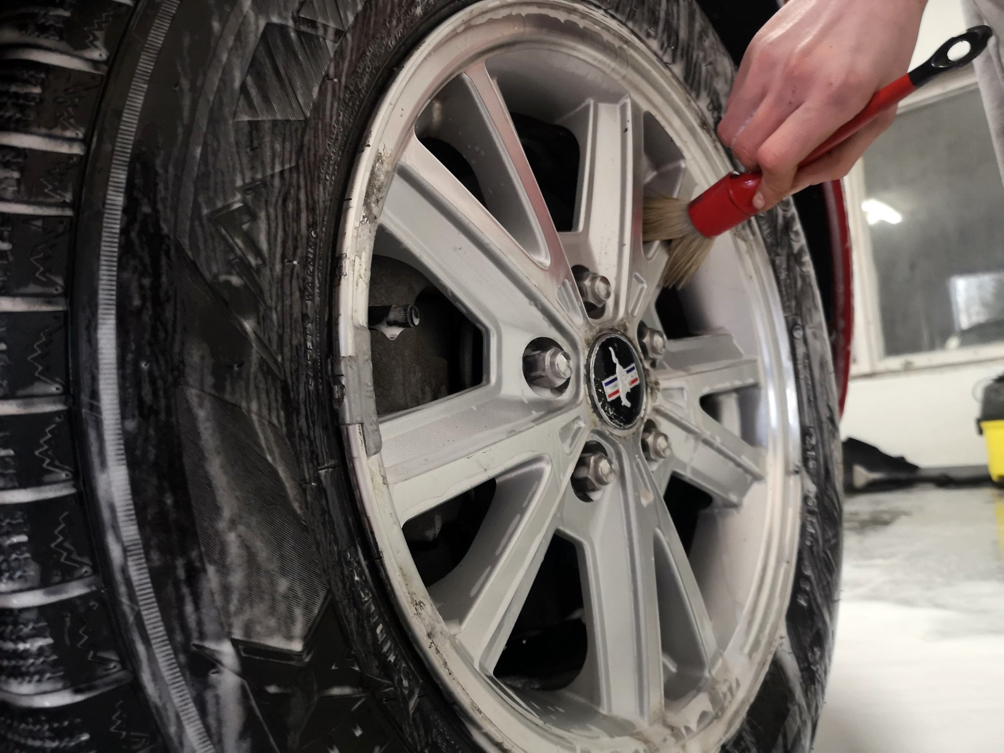 Szczotkowanie felgi aluminiowej samochodu z bliska, widoczna piana na oponie i czerwona szczotka w dłoni.