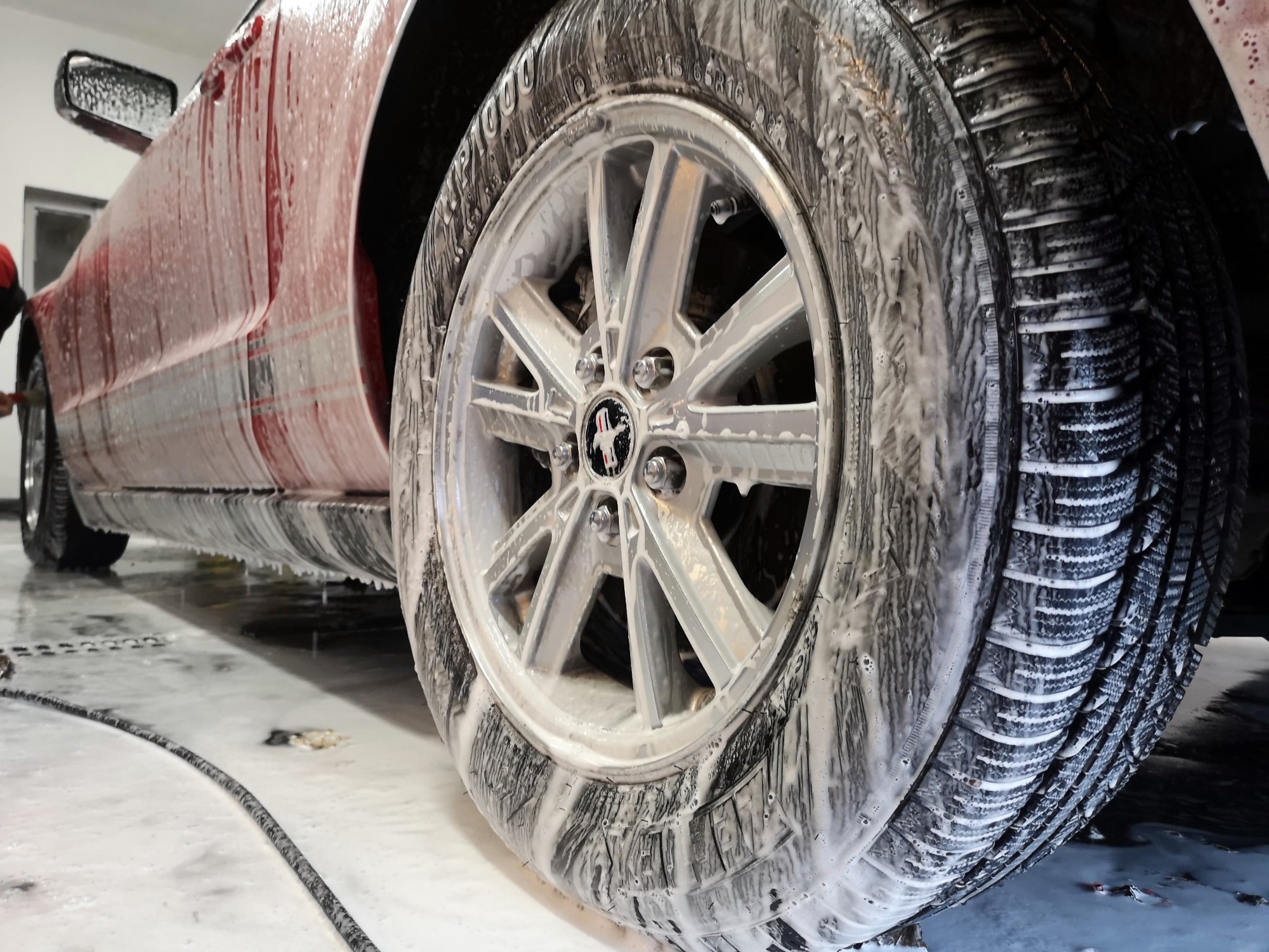 Czerwony samochód w trakcie mycia, z bliska widoczne koło pokryte pianą, felga z logo konia.