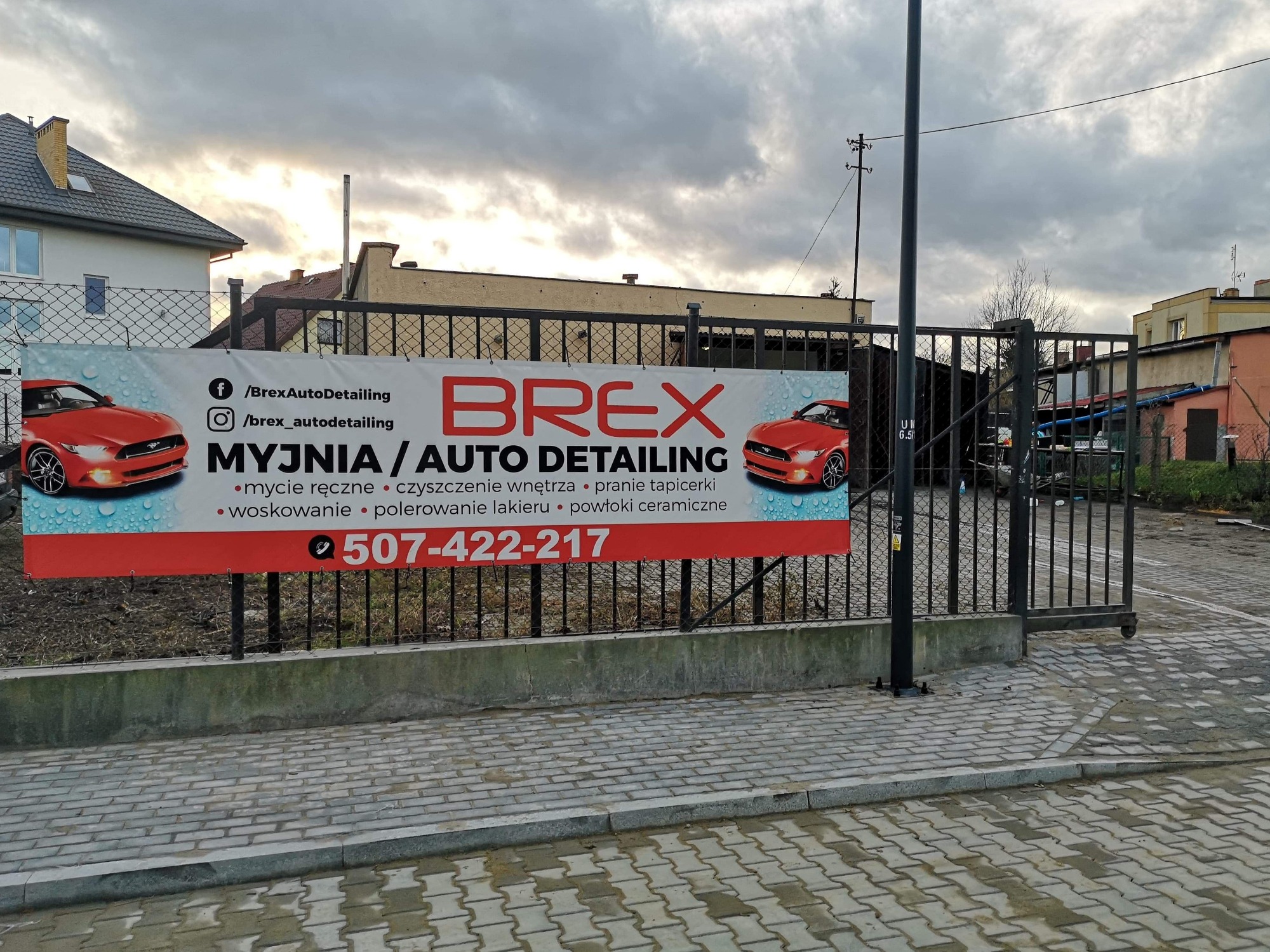 Baner reklamowy myjni samochodowej Brex Auto Detailing z czerwonym sportowym autem, umieszczony na ogrodzeniu w pochmurny dzień.