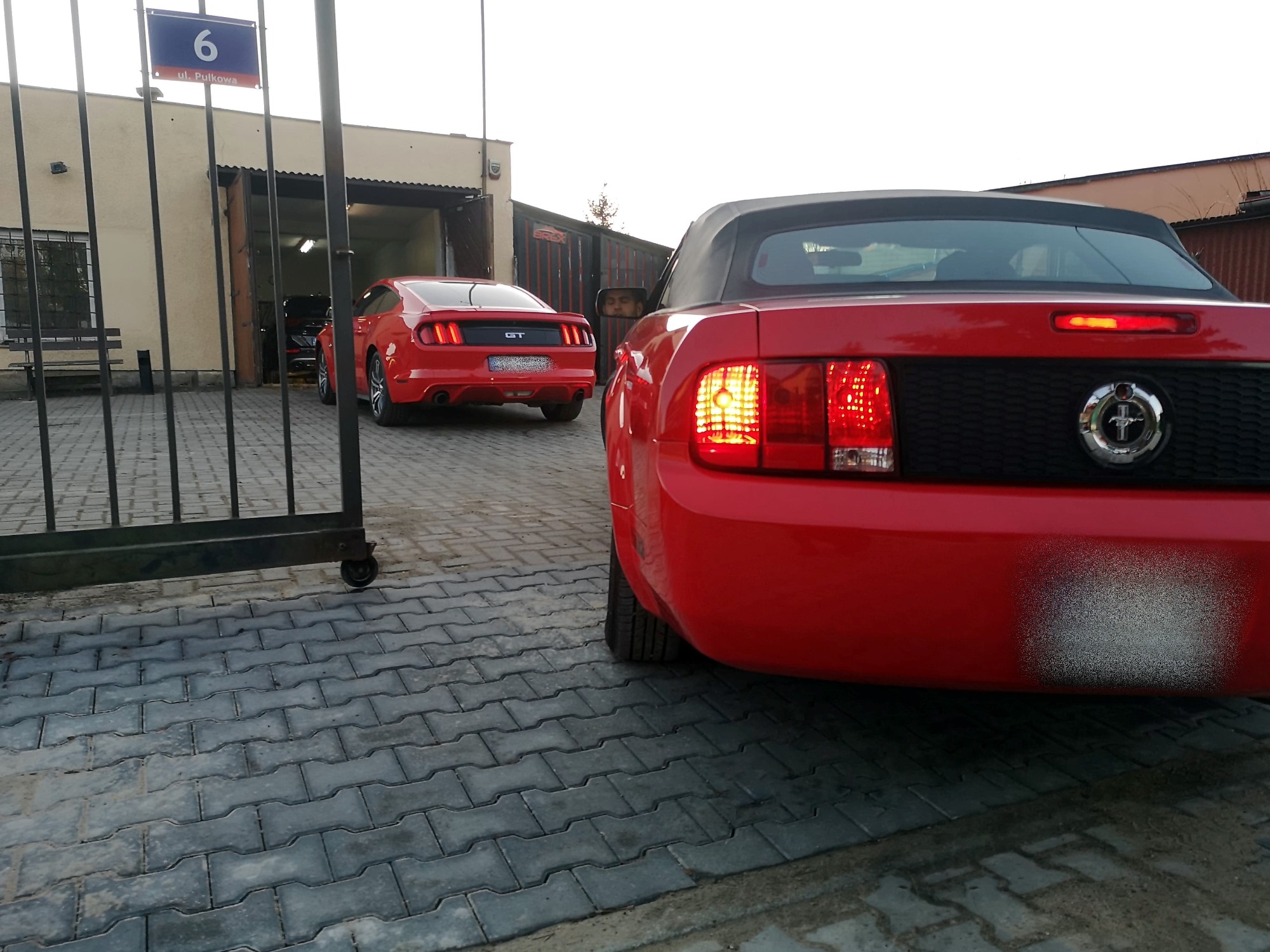 Dwa czerwone samochody typu kabriolet marki Ford Mustang zaparkowane przed warsztatem, widok z tyłu, z widoczną tablicą adresową 'ul. Pułkowa 6'.