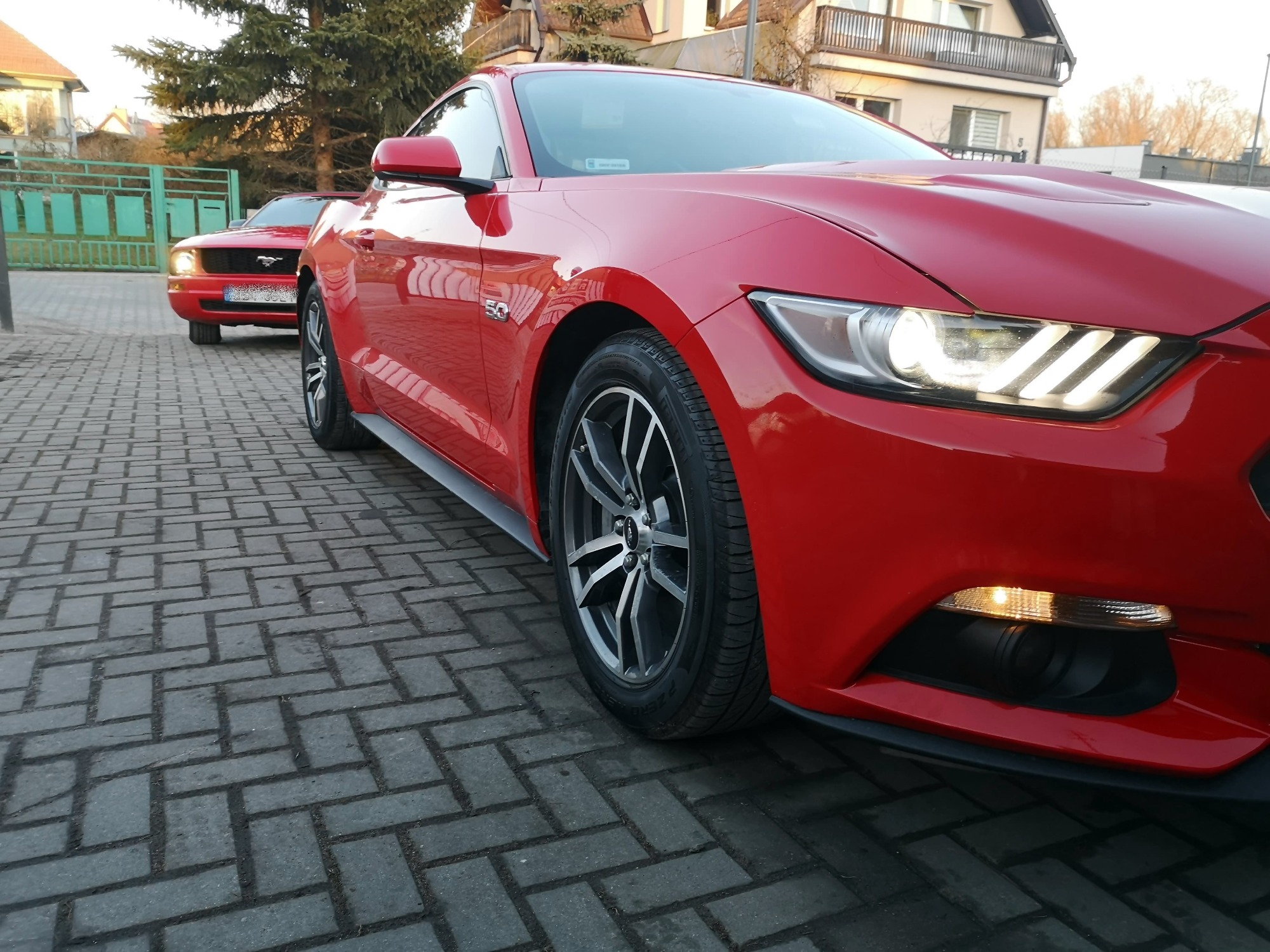 Dwa czerwone samochody Ford Mustang zaparkowane na brukowanej drodze przed domem, z bliska widoczny model z 2015 roku z oznaczeniem '5.0' na błotniku.