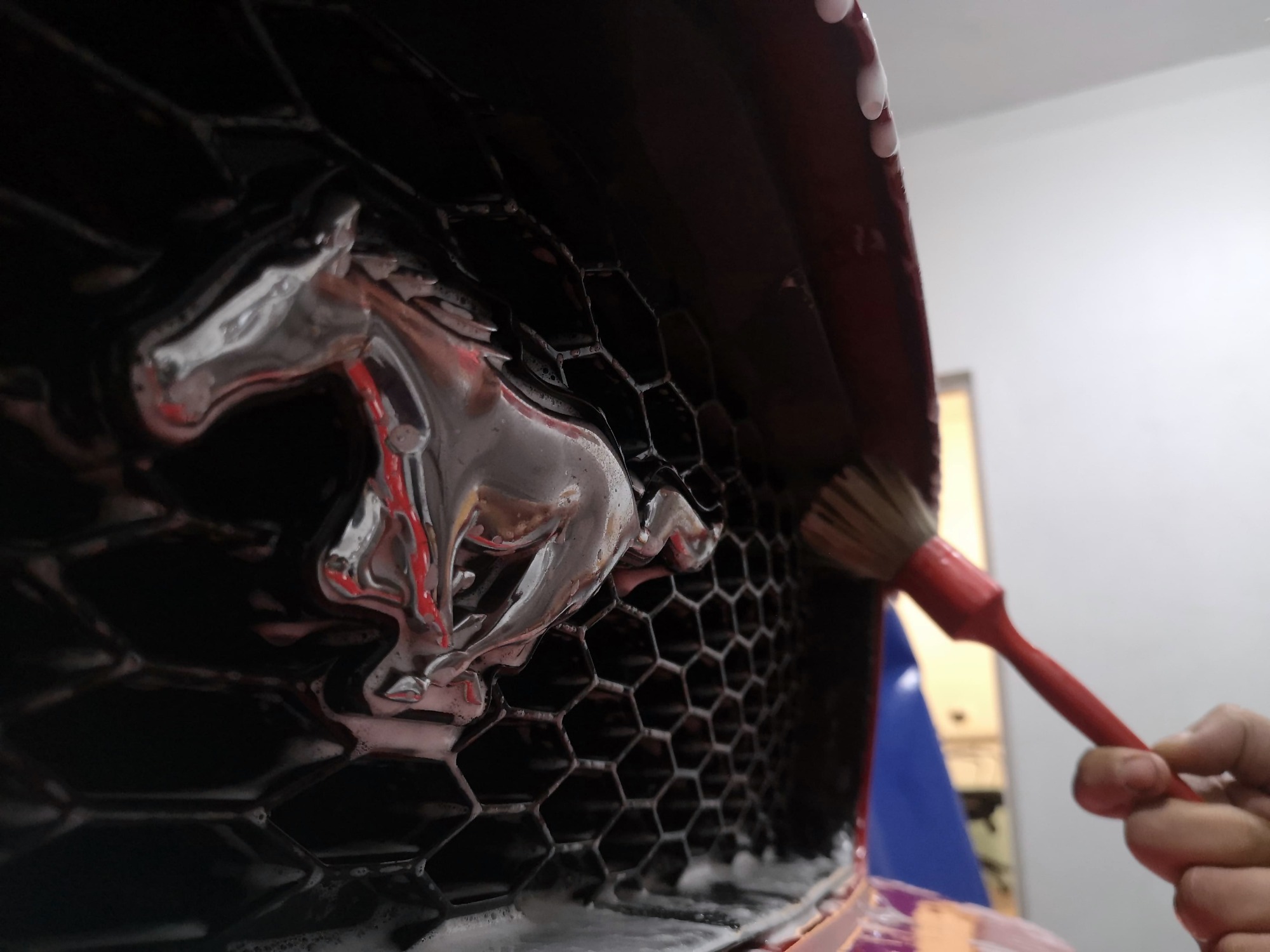 Czyszczenie grilla samochodu z chromowanym logo konia za pomocą pędzelka z czerwonym trzonkiem i piany aktywnej.