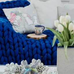 Stylowa kompozycja wielkanocna z niebieskim, grubym kocem, poduszką z motywem królików, kubkiem na drewnianym plastrze, białymi tulipanami w ceramicznym wazonie i wiankiem z króliczkami.