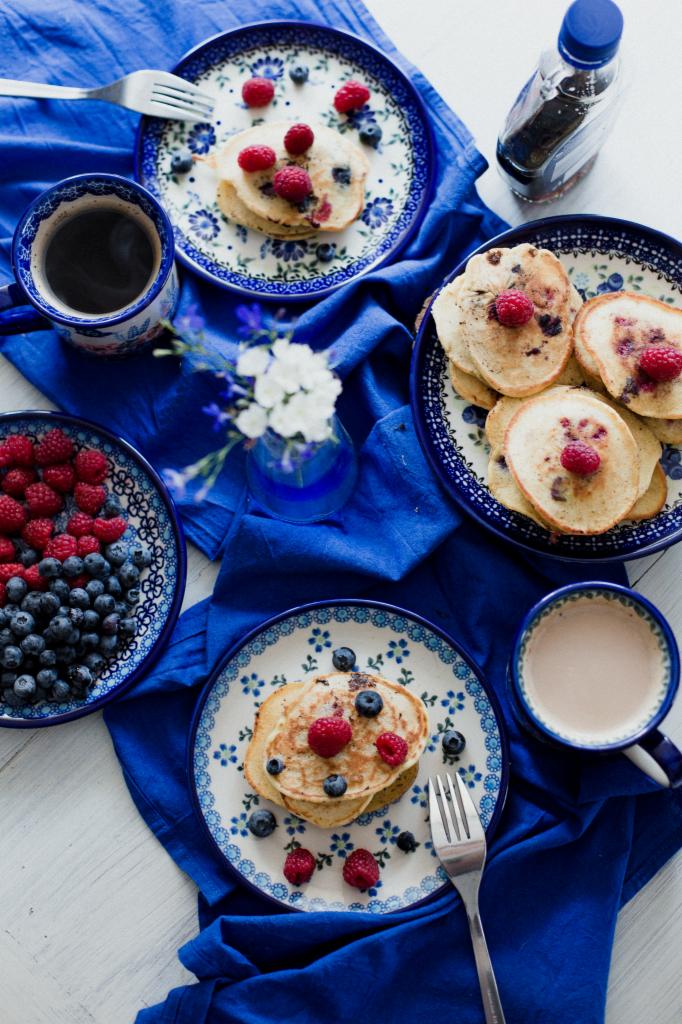 Stylowe zdjęcia produktowe: Pancakes z malinami i borówkami na talerzach w niebieskie wzory, kawa w filiżance, owoce w miseczce i mały wazonik z kwiatami, wszystko ułożone na niebieskiej tkaninie