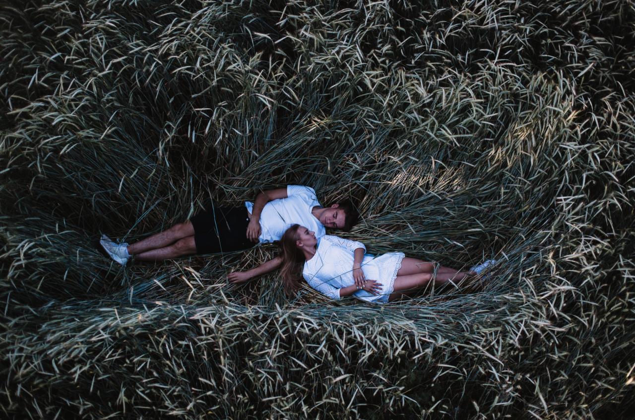 Para leży w trawie, widok z góry, on w białym t-shircie i czarnych spodenkach, ona w białej sukience z koronkowymi detalami.