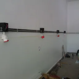 Montaż instalacji elektrycznej w nowym budynku, widoczne czarne rury prowadzące kable, puszki instalacyjne na ścianie oraz tymczasowe oświetlenie budowlane z czerwonymi kloszami, w tle rozmazana...