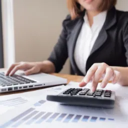 Kobieta w czarnej marynarce kalkuluje dane na kalkulatorze i laptopie, analizując wykresy.