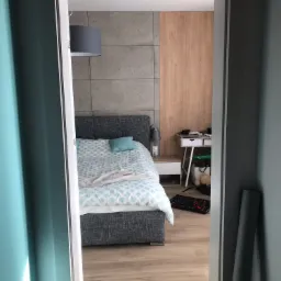 Widok z korytarza na sypialnię z łóżkiem w odcieniach szarości i błękitu, betonową ścianą i drewnianym panelem, biurkiem z akcesoriami oraz narzędziami na podłodze.