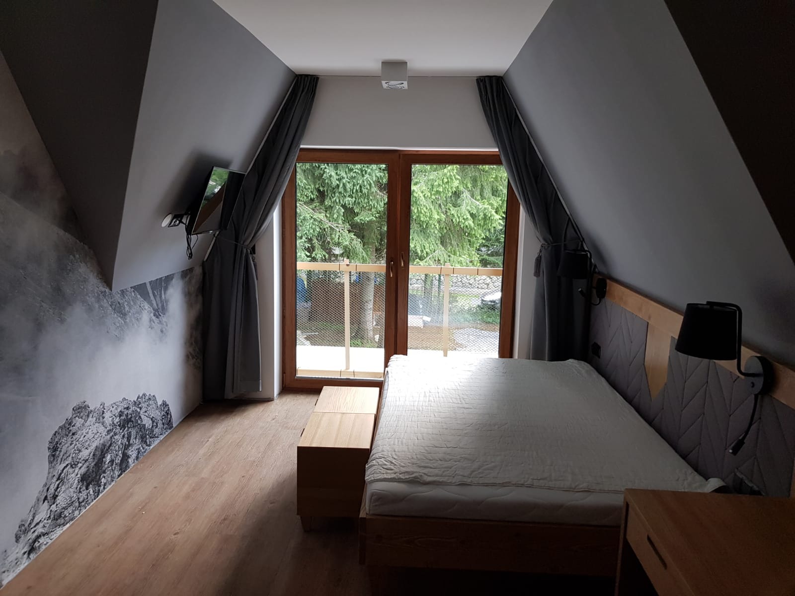 Pokój na poddaszu w stylu górskim z łóżkiem, drewnianą podłogą, fototapetą z motywem górskim i widokiem na las z balkonu.