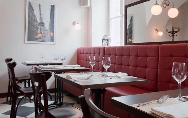 Wnętrze bistro z czerwonymi tapicerowanymi lożami, nakrytymi stołami, szklankami i sztućcami, na ścianie grafika z wieżą Eiffla.
