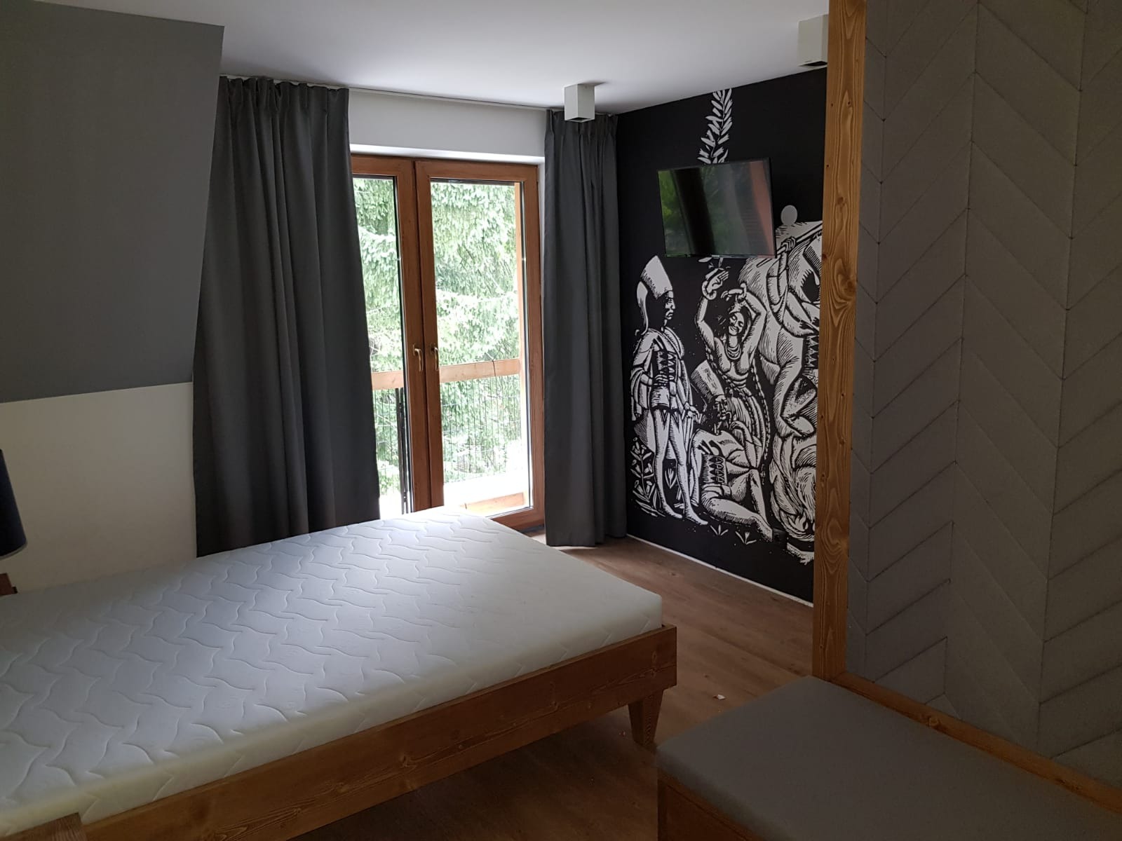 Pokój w apartamencie z drewnianym łóżkiem, balkonem z widokiem na las i ścianą z czarno-białym muralem przedstawiającym postacie w stylu historycznym, nad którym wisi telewizor.
