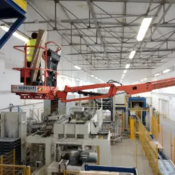 Pracownik w pomarańczowym podnośniku koszowym z logo AA Herkules, wykonujący prace na wysokości w hali produkcyjnej z dużą maszyną przemysłową i paletami w tle.