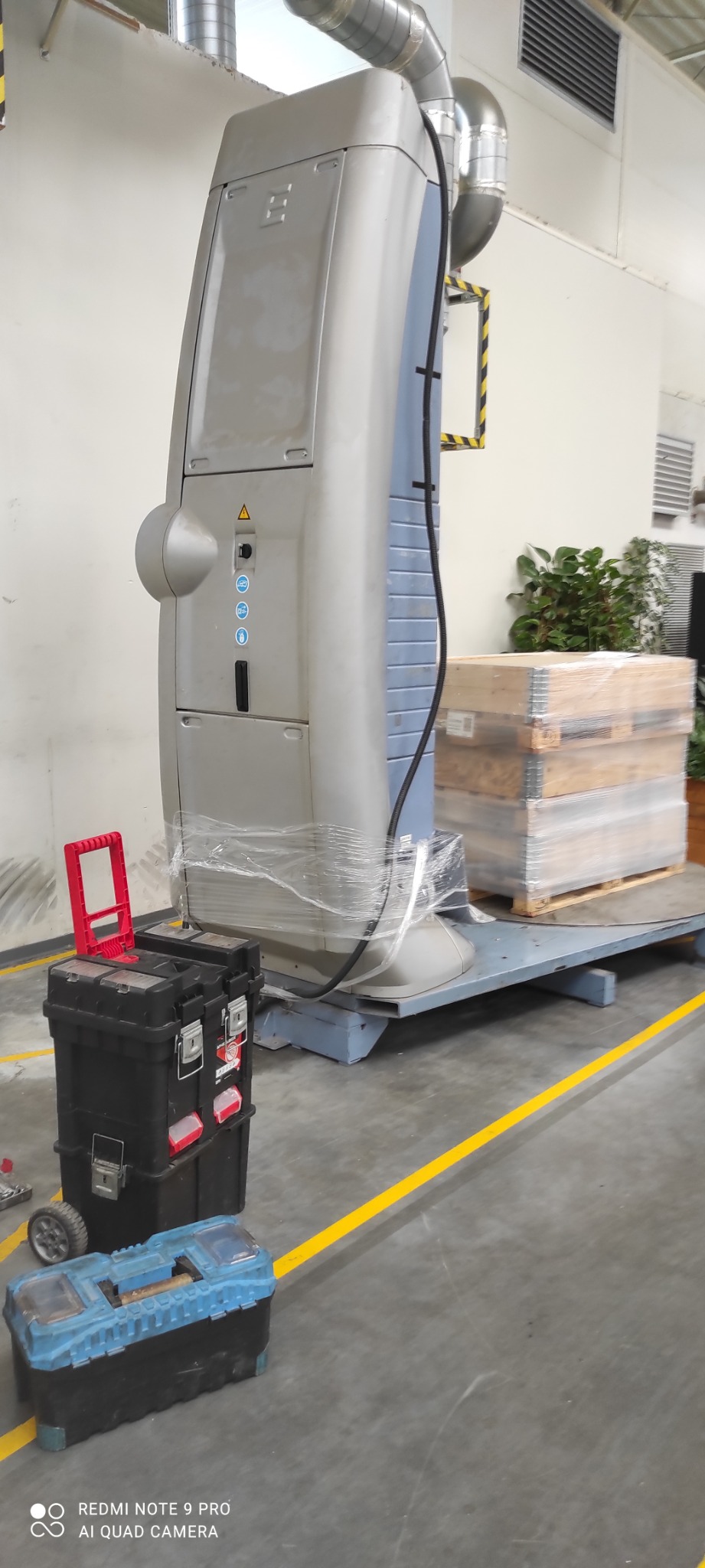 Duża, szara maszyna przemysłowa owinięta folią stretch, stojąca na metalowej platformie w hali. Obok skrzynki narzędziowe: czarna z czerwoną rączką i niebieska. W tle widoczne palety z drewnianymi...